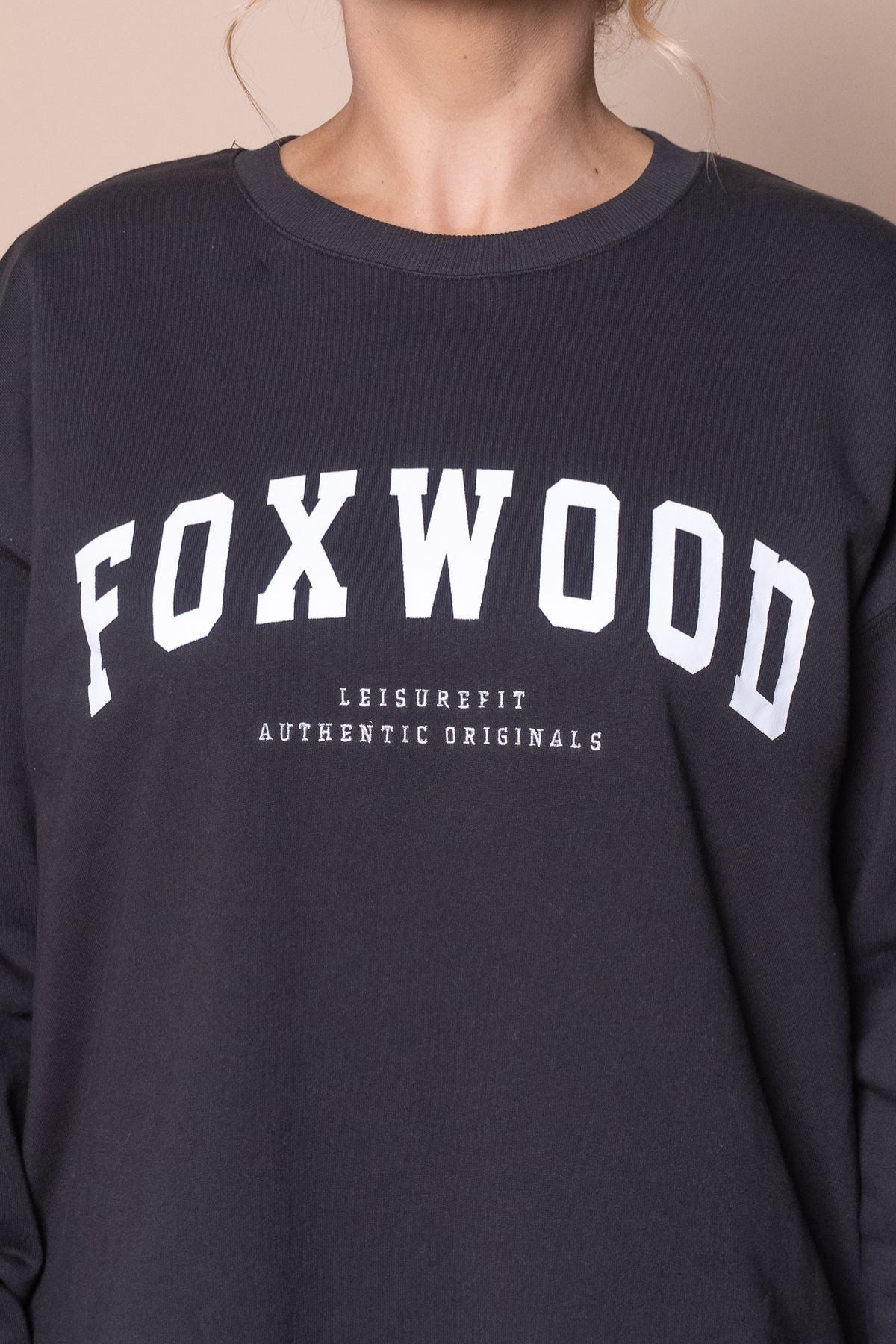 A person wears a black Foxwood Interval Crew sweatshirt with "FOXWOOD" in large white letters and "LEISUREFIT AUTHENTIC ORIGINALS" below. This relaxed-fit everyday essential is shown without the person's face visible.