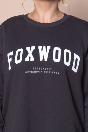 A person wears a black Foxwood Interval Crew sweatshirt with "FOXWOOD" in large white letters and "LEISUREFIT AUTHENTIC ORIGINALS" below. This relaxed-fit everyday essential is shown without the person's face visible.