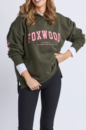 A woman with wavy hair wears the oversized olive-green Foxwood Interval Hoodie, featuring pink lettering, styled with black leggings. She stands smiling with one hand on her hip—showcasing this versatile layer for casual outings.