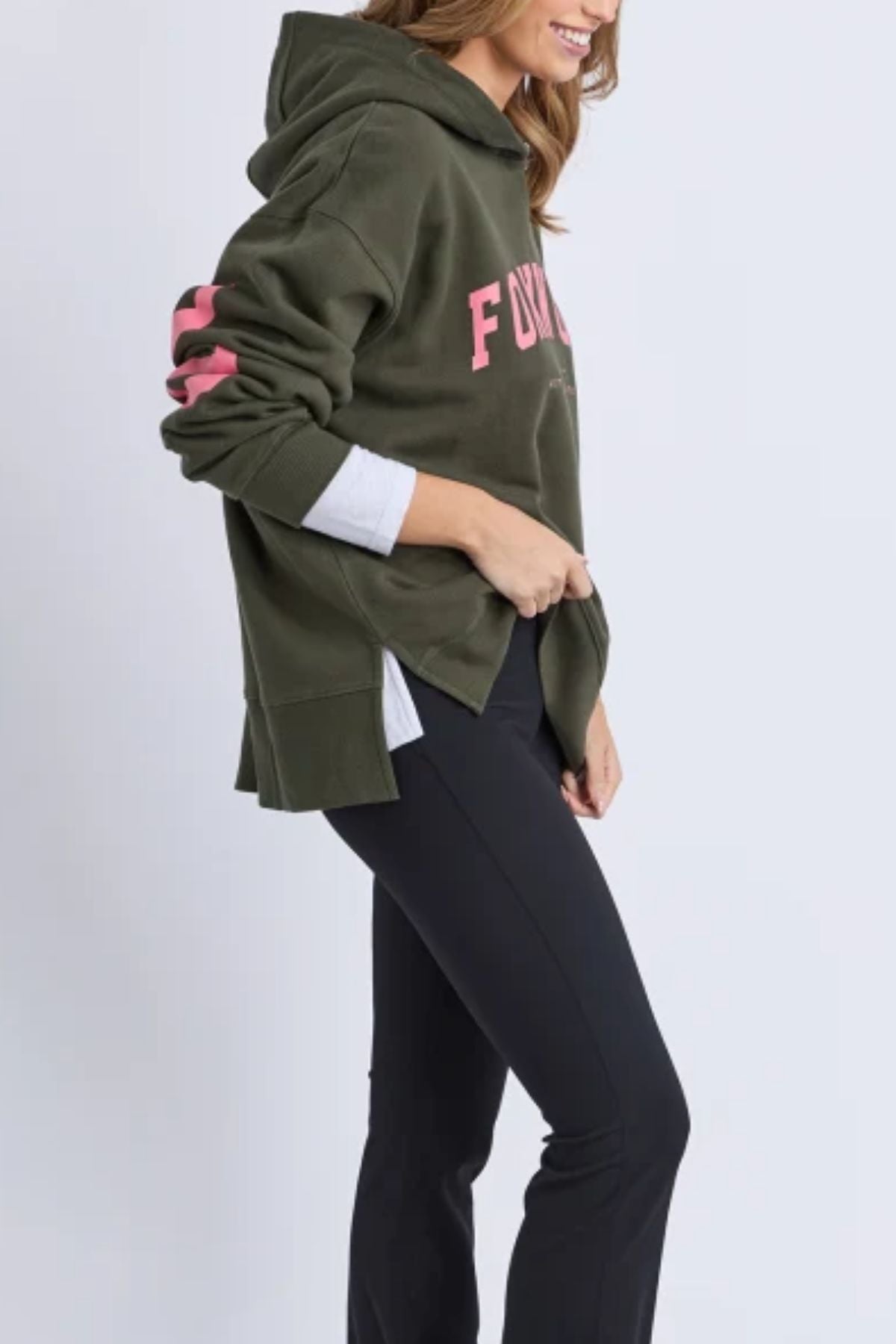 A person wearing the Foxwood Interval Hoodie with pink lettering, a white shirt underneath, and black pants stands sideways, smiling and casually holding the hem—a stylish choice for everyday wear.