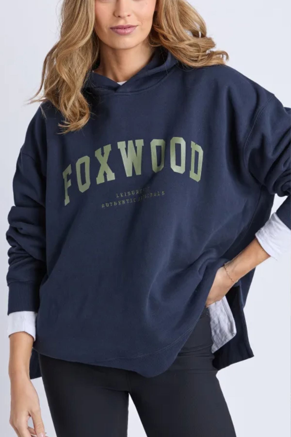 A woman models the Foxwood Interval Hoodie in green, featuring a prominent logo, layered over a white shirt and black leggings. Her relaxed look is enhanced by wavy light brown hair, softly framing her face as she stands with one hand in her pocket.