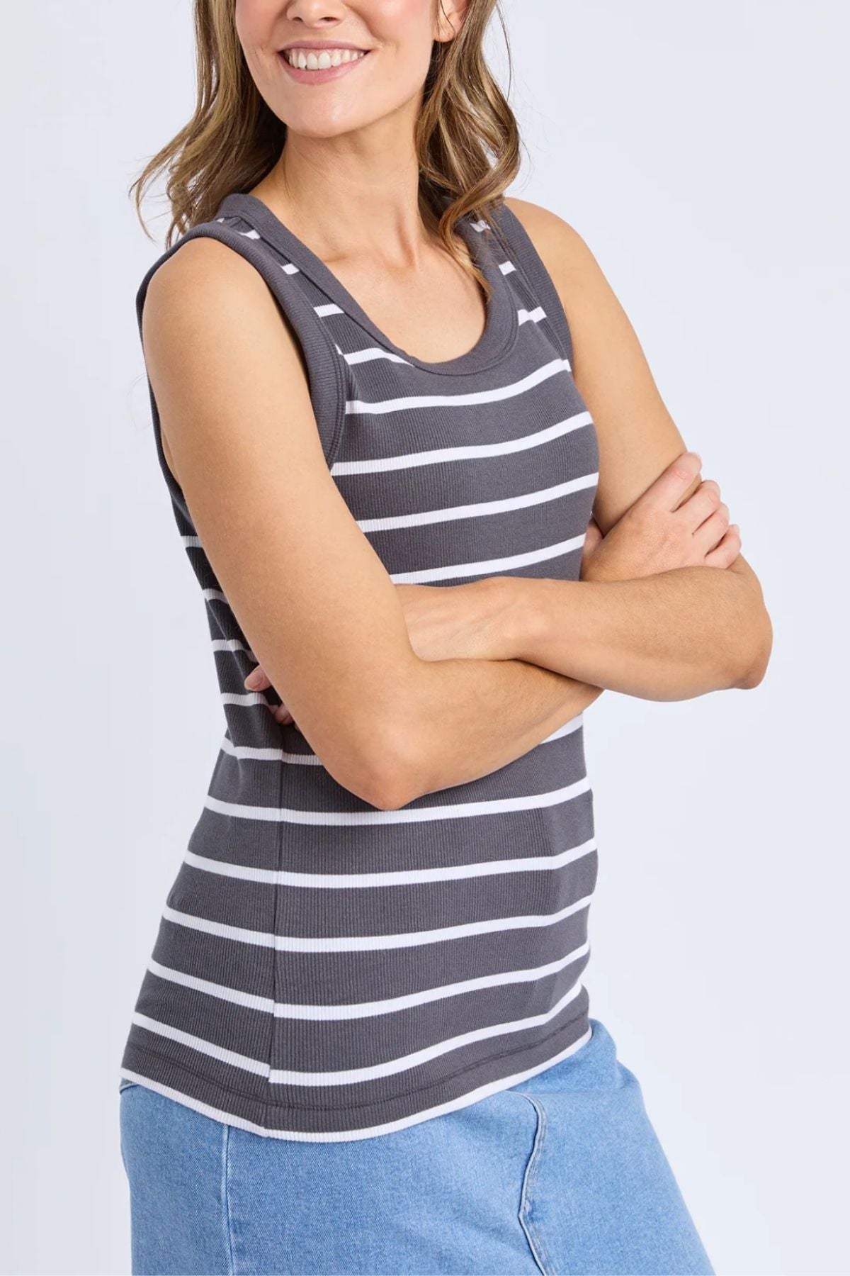 A woman with wavy brown hair smiles with arms crossed, wearing the Foxwood Iris Stripe Tank—a relaxed fit gray tank with white stripes—paired with a light blue denim skirt against a plain background.