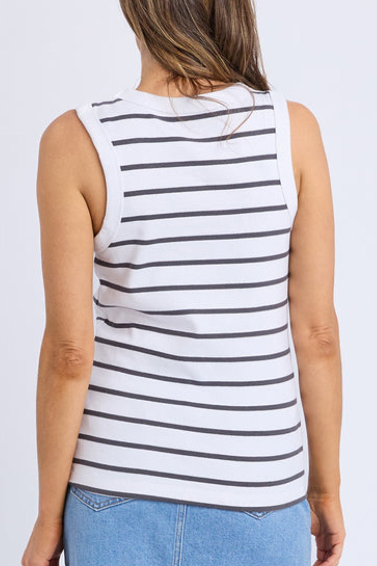 A person with long hair is seen from the back wearing a sleeveless white Foxwood Iris Stripe Tank with horizontal black stripes, paired with light blue jeans, against a plain background.
