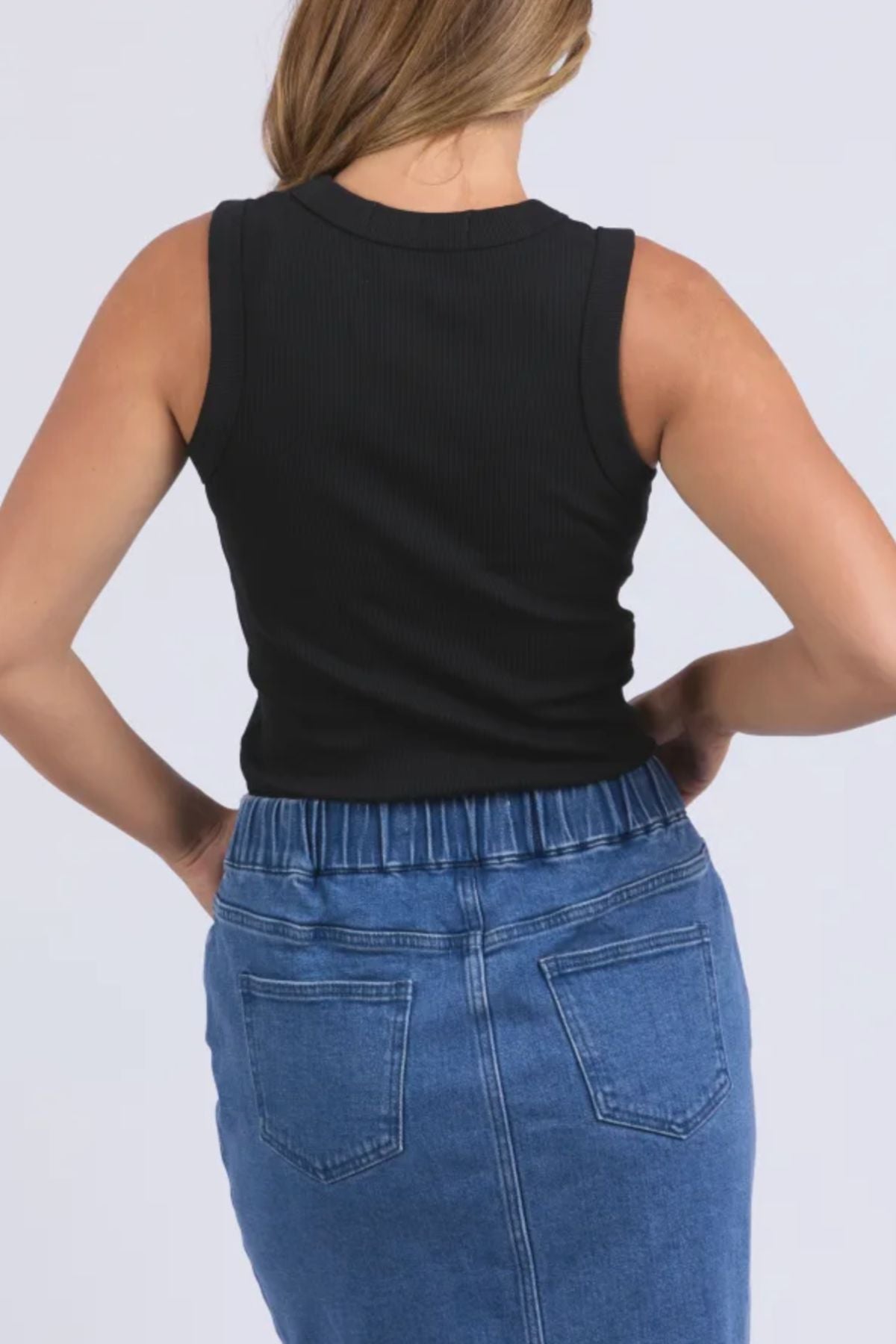A person with light brown hair wears the sleeveless black Foxwood Iris Tank paired with a blue denim skirt, photographed from behind against a plain light background.