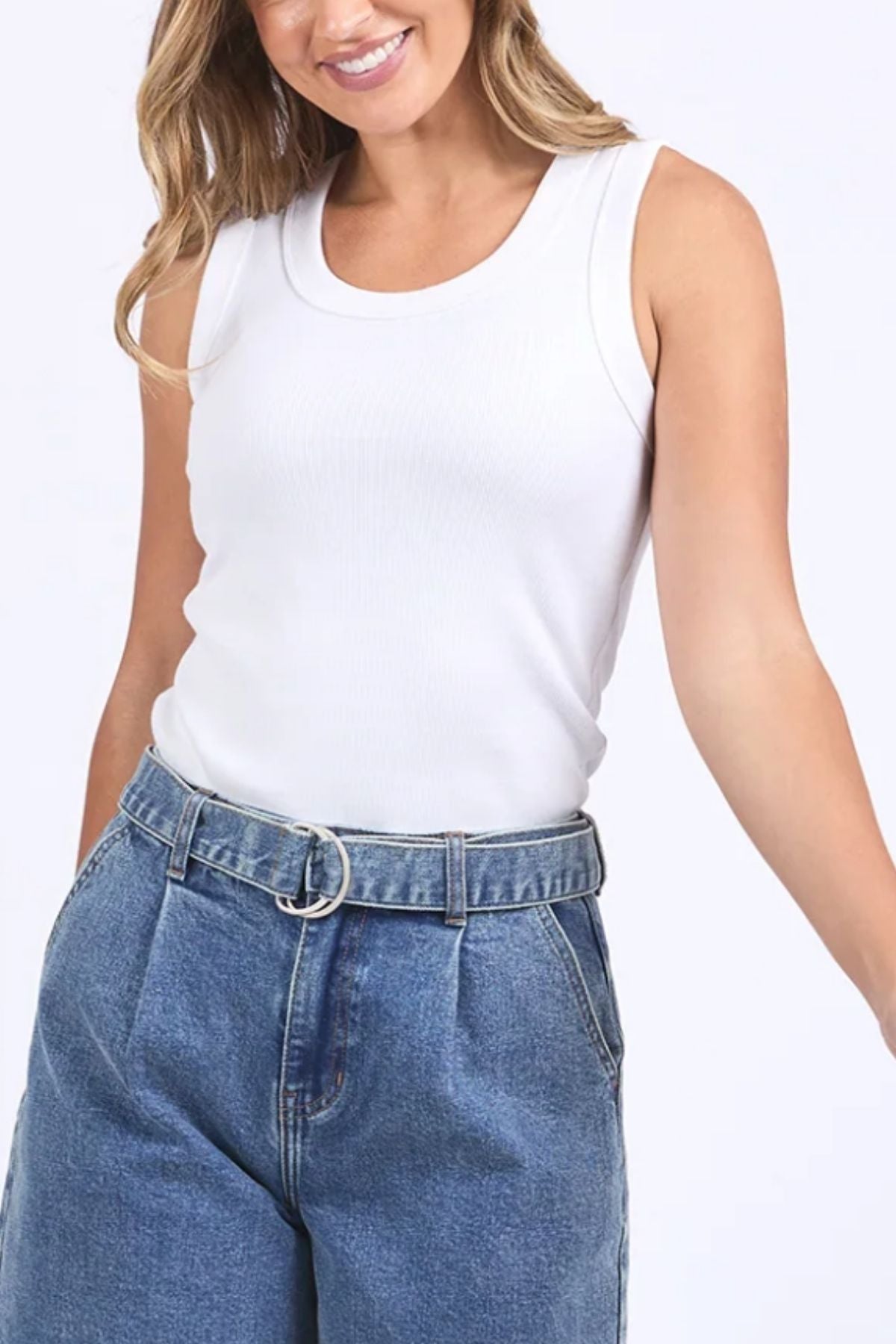 A woman smiles wearing a sleeveless Foxwood Iris Tank in white ribbed stretch fabric, tucked into high-waisted blue jeans with a circular belt buckle, her left arm slightly outstretched.
