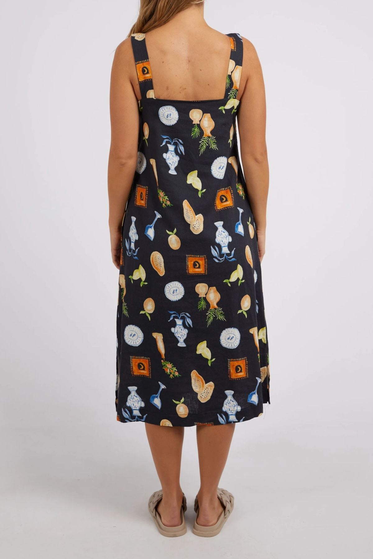 A woman is shown from the back in a sleeveless Foxwood Italian Summer Dress that reaches the knees. The dress features a lively food-themed pattern with fruits and vegetables set against a simple background. Made from soft linen, it is ideal for sunny occasions. She completes her relaxed look with slippers.