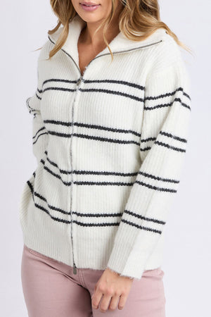 A woman models the Foxwood Journey Zip Knit, a white sweater with black stripes and zip-up front, paired with pink pants. She stands against a plain background with one hand in her pocket, displaying its relaxed fit.