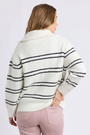 A woman stands with her back to the camera, wearing a white Foxwood Journey Zip Knit with black stripes and light pink pants. The sweater’s soft, ribbed knit and relaxed fit offer comfort as she rests her right hand on her hip.