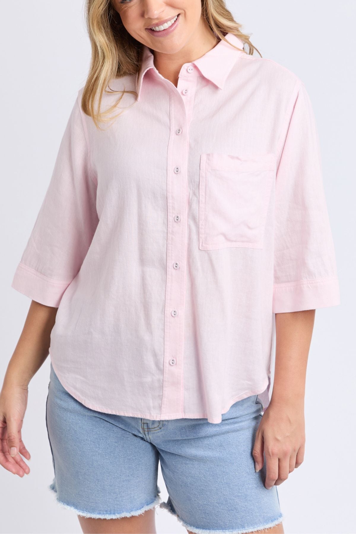 A woman wears the Foxwood Leah Shirt, a light pink linen top with a relaxed fit, chest pocket, and 3/4 sleeves, paired with light blue denim shorts, smiling against a plain background.