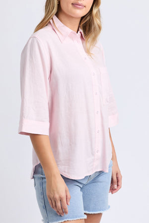 A woman wears the Foxwood Leah Shirt, a relaxed fit linen top in light pink with 3/4 sleeves, paired with light blue denim shorts against a plain background.