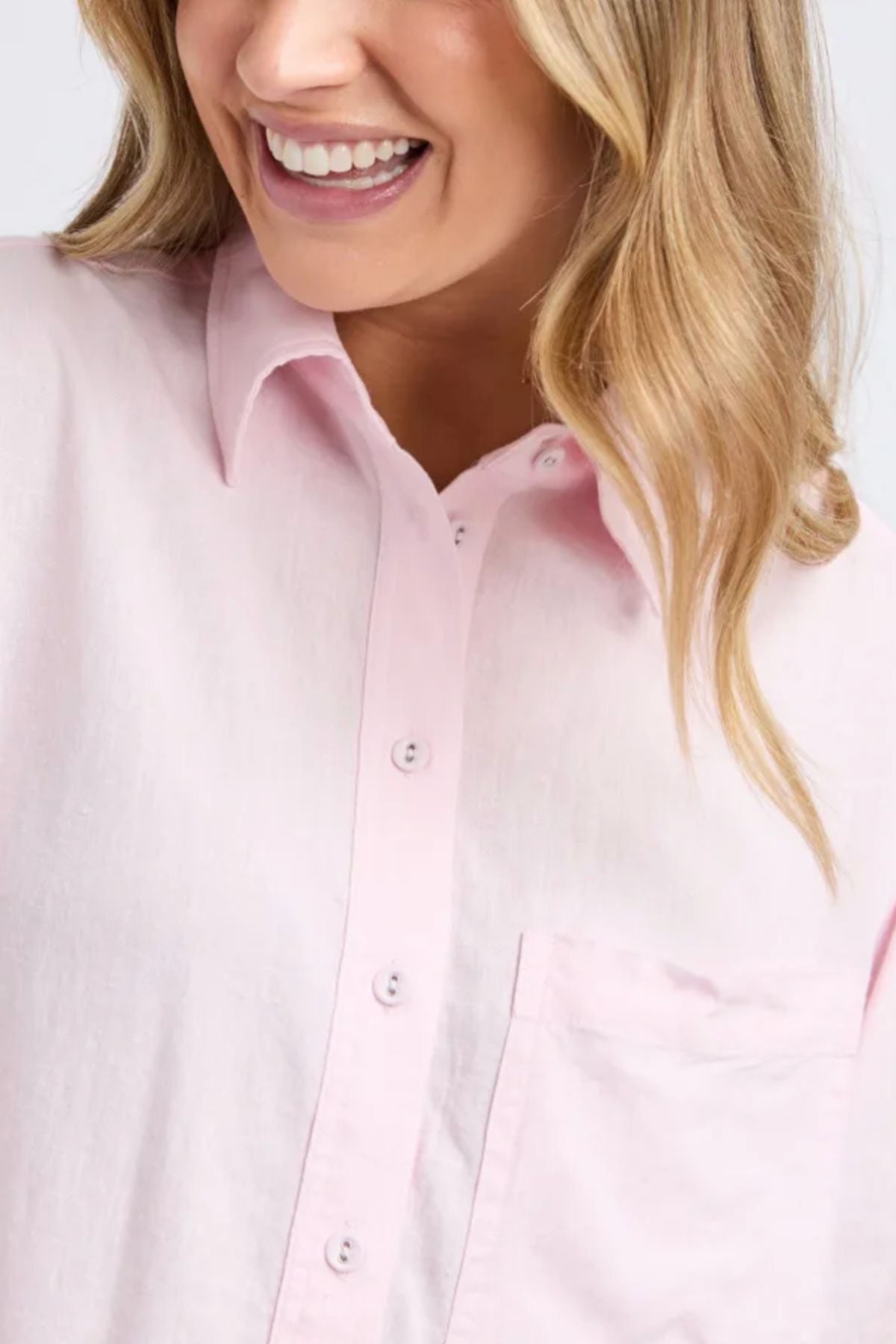 A woman smiles in the Foxwood Leah Shirt, a pale pink linen top featuring 3/4 sleeves and a chest pocket. Only her lower face, some hair, and upper torso are shown, highlighting the shirt’s relaxed fit.