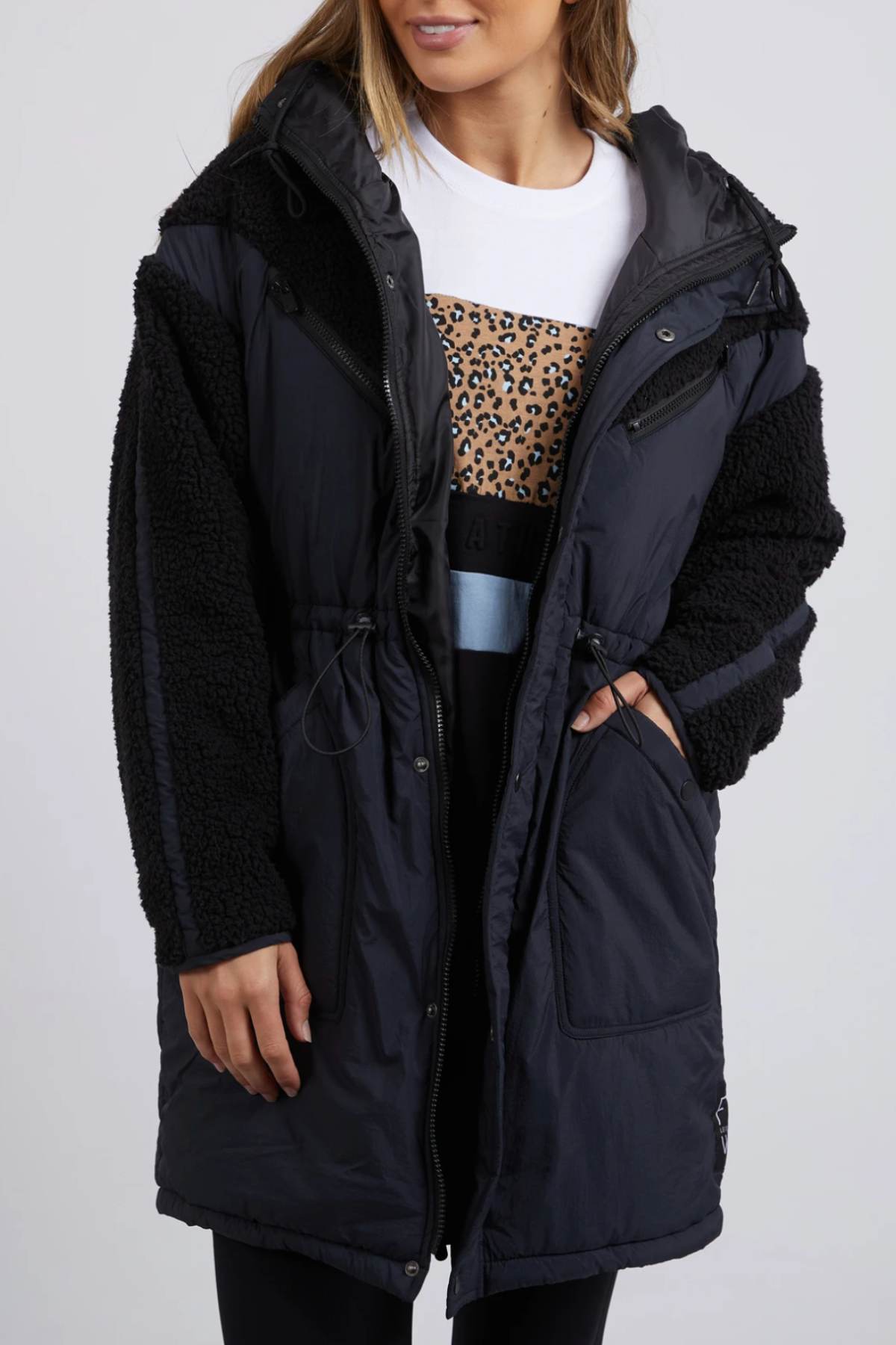 The woman wears a Foxwood Longline Anorak with faux shearling sleeves, hiding her leopard-print sweater and black pants. Her hand in her pocket, she stands against a plain background, prepared for adventure in her durable nylon polyester blend ensemble.