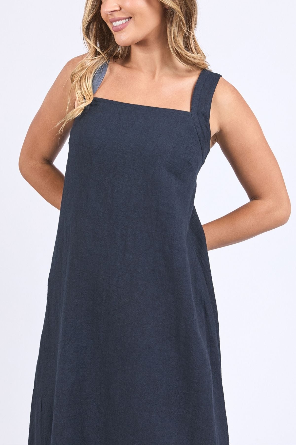 A woman with long, wavy blonde hair is wearing the Foxwood Lourdes Dress—a sleeveless, loose-fitting navy blue linen dress with wide straps and a square neckline—while smiling against a plain white background.