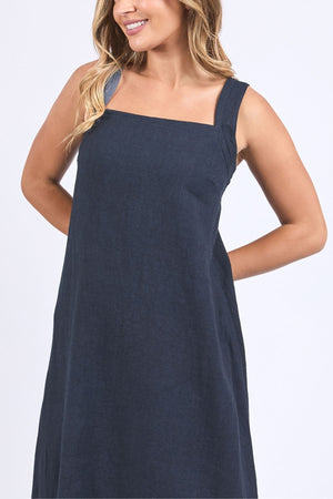 A woman with long, wavy blonde hair is wearing the Foxwood Lourdes Dress—a sleeveless, loose-fitting navy blue linen dress with wide straps and a square neckline—while smiling against a plain white background.
