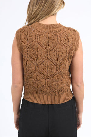 A person shown from the back wears the sleeveless Foxwood Luna Knit Vest in brown, featuring a leaf-like pointelle pattern and ribbed edges, paired with high-waisted black pants.