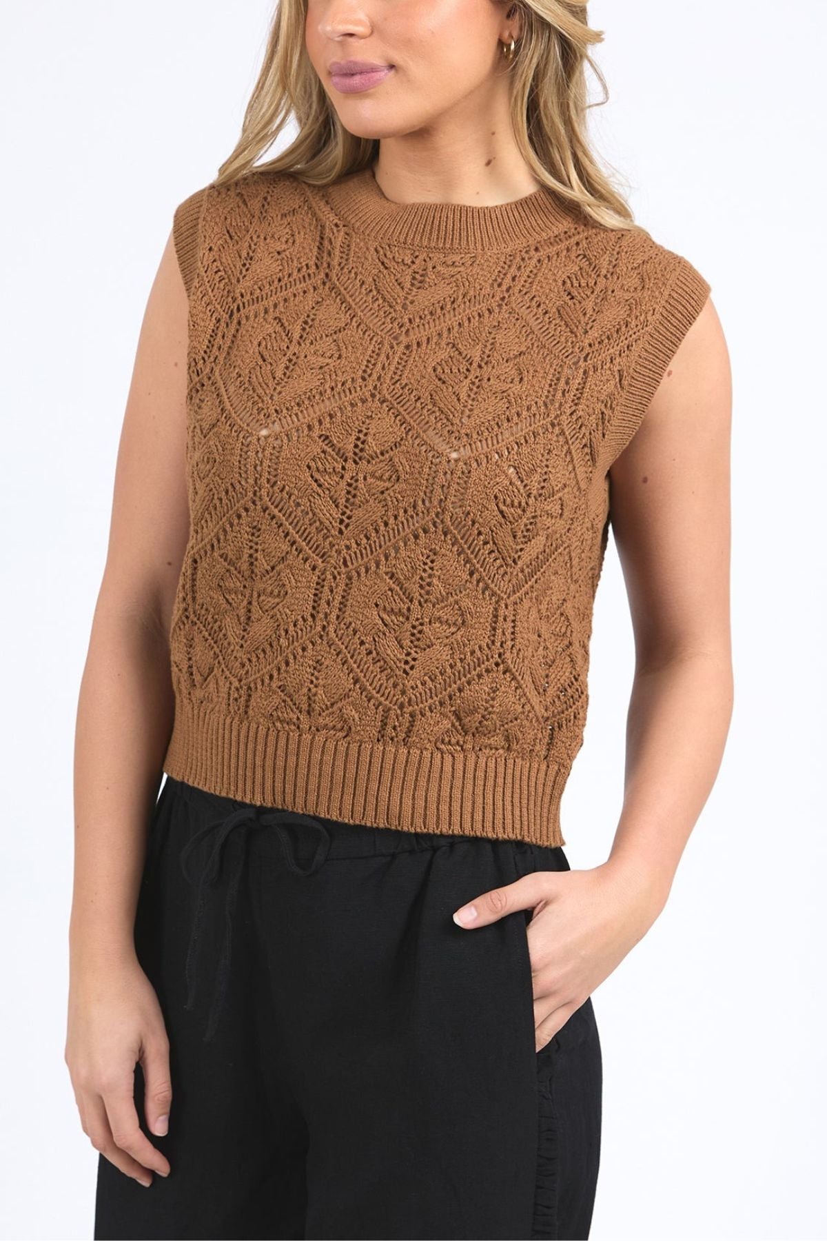 Woman wears the Foxwood Luna Knit Vest—a sleeveless, brown pointelle knit top with a geometric pattern—paired with high-waisted black pants. The photo focuses on her upper body; her head is partially out of frame.