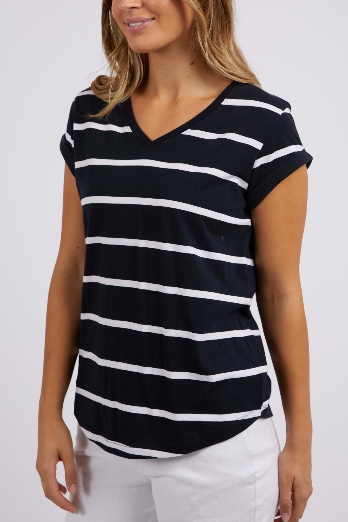 A woman is wearing the Foxwood Manly Stripe Vee Tee, a black Pima cotton jersey T-shirt with white yarn-dyed stripes, featuring a vee neckline and short sleeves. She is also wearing white pants against a plain and neutral background.