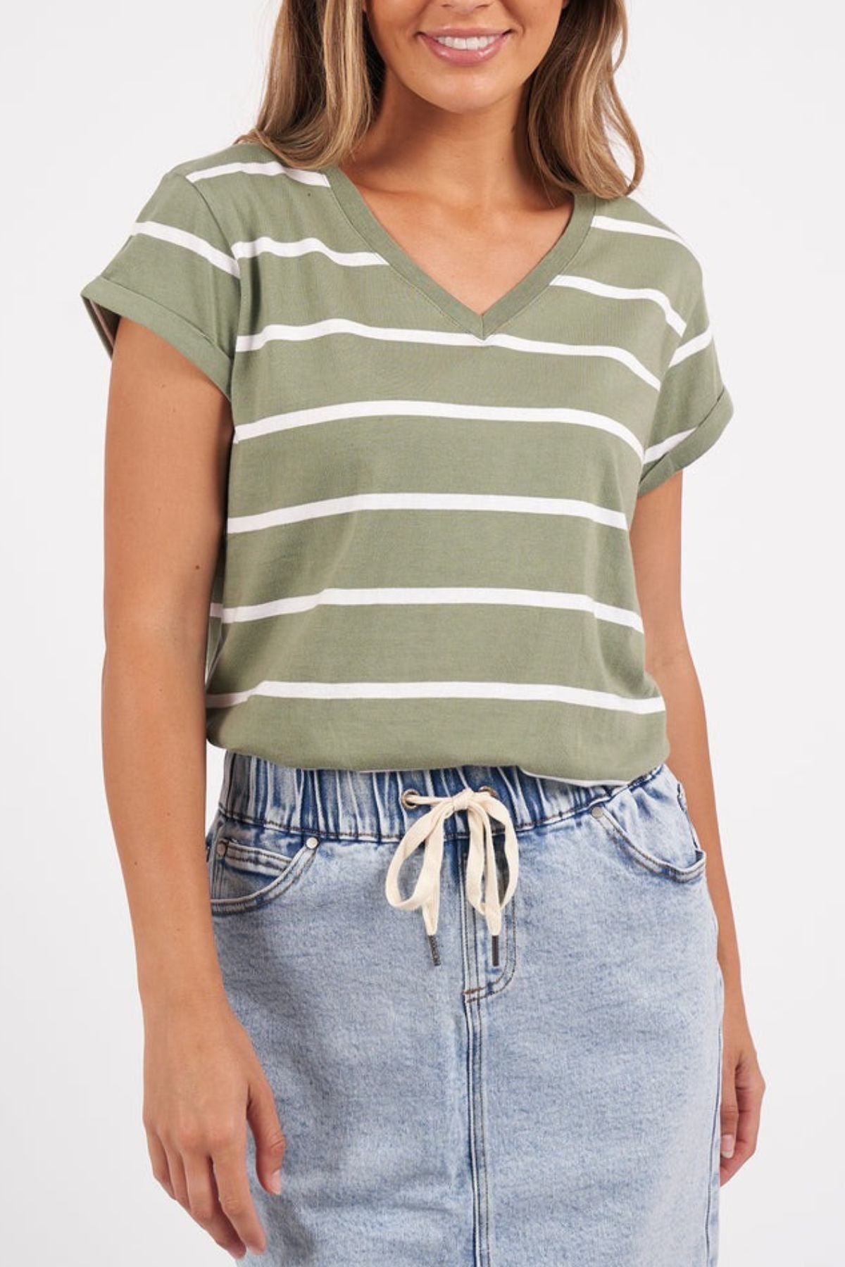 A person is dressed in the Foxwood Manly Stripe Vee Tee, a chic tee with green and white stripes made from Pima cotton jersey, paired with a light blue denim skirt that has a white drawstring. The plain white background enhances the scene's simplicity.