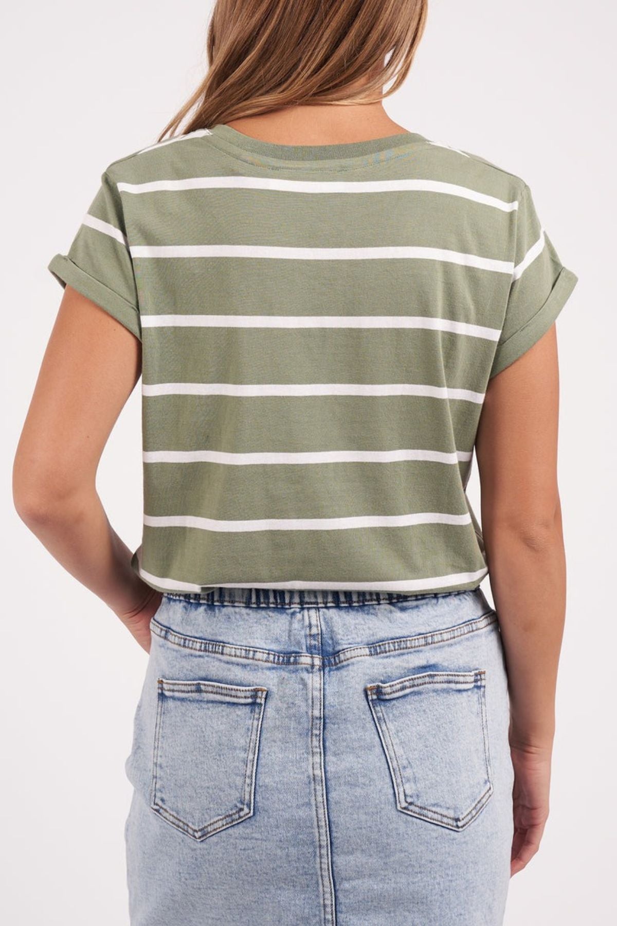 A person wearing the Foxwood Manly Stripe Vee Tee, featuring green and white stripes in soft Pima cotton jersey, stands facing away from the camera. Complementing their outfit is a light blue denim skirt, set against a plain white background to accentuate the tee's subtle elegance.