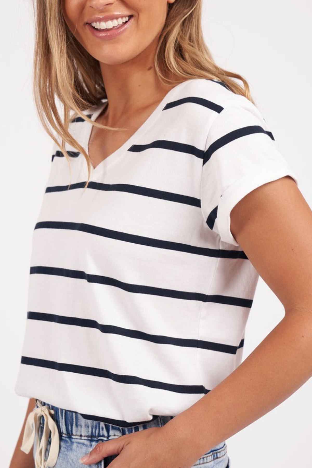 A woman in a Foxwood Manly Stripe Vee Tee smiles, posing with one arm on her waist. The vee neckline complements her long blonde hair, and the outfit includes casual blue jeans. The background is plain white.