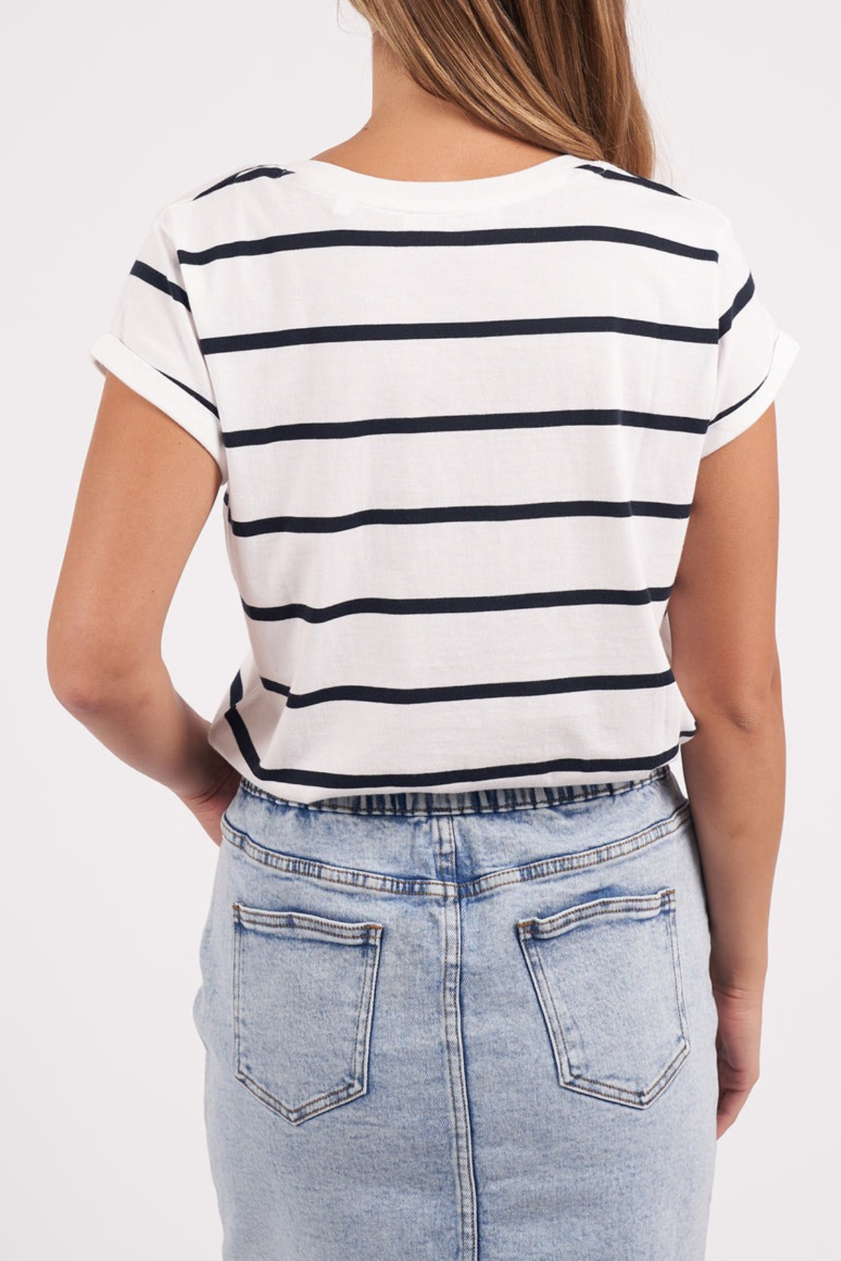 A person wearing the Foxwood Manly Stripe Vee Tee, a white and black yarn dyed stripe short-sleeve shirt made of comfy Pima cotton jersey, and a blue denim skirt with two back pockets is facing away from the camera. The skirt stands out against the plain white background.