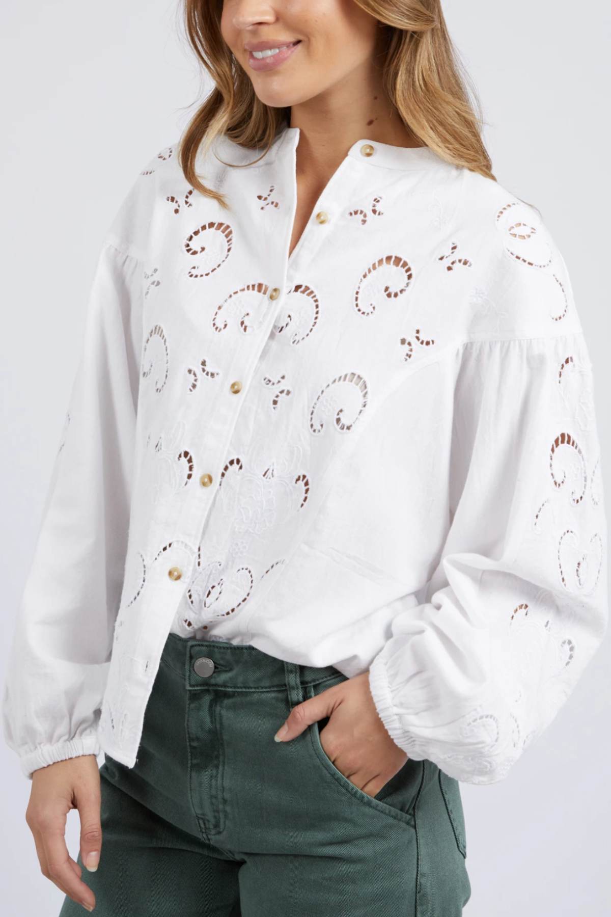 A woman smiles with her hand in her pocket, wearing the Foxwood Marlow Blouse—a white, long-sleeve cotton button-up with eyelet-style embroidery—paired with high-waisted green pants.