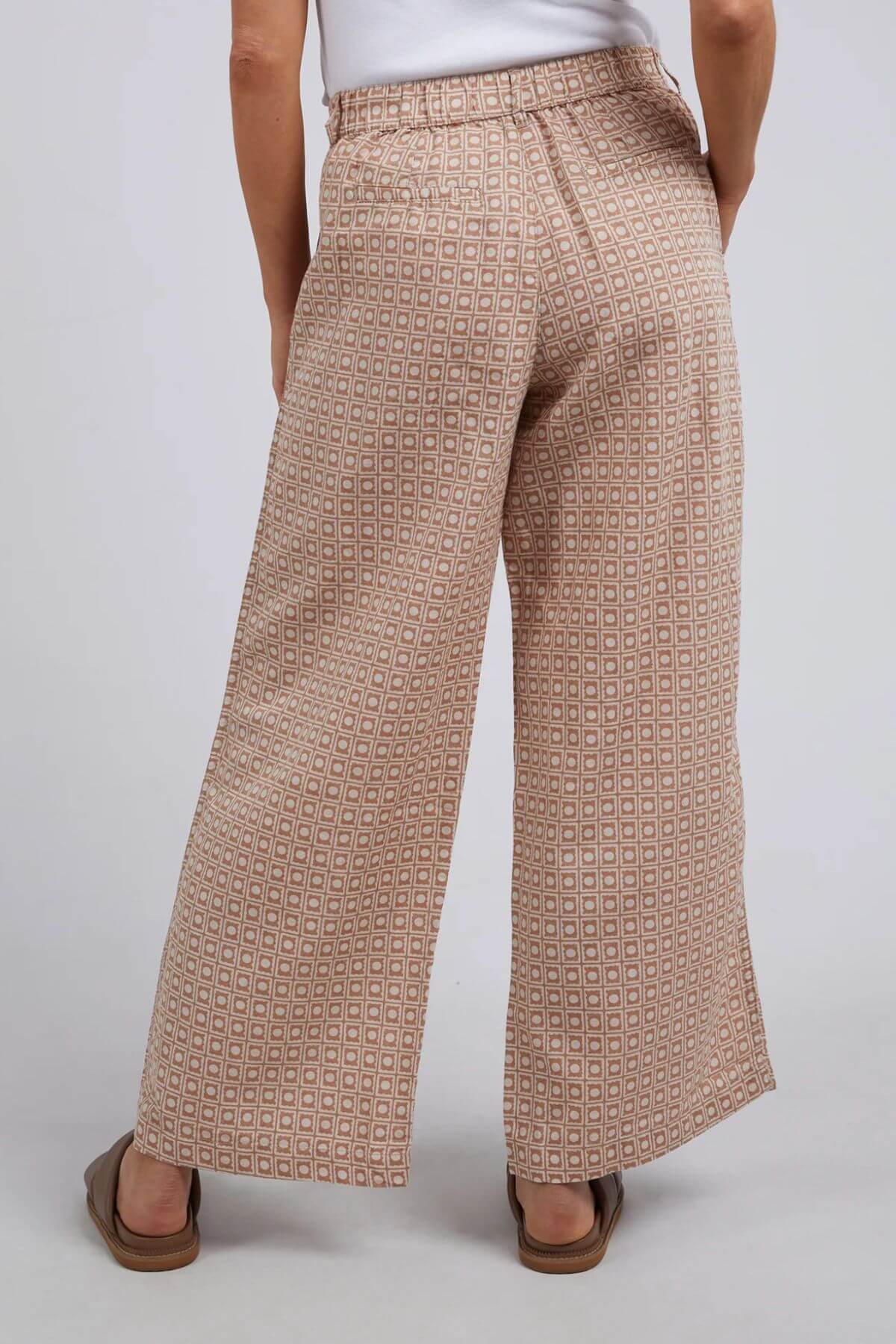 A person wearing Foxwood Maya Geo Pant, made from a linen/cotton blend, stands facing away. These wide-leg, light brown pants feature a small geometric pattern and an elastic waistband. Styled with a white top and brown sandals against a light gray backdrop.