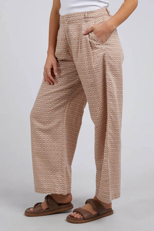 Wearing the Foxwood Maya Geo Pant in a beige color, a person showcases style and comfort with its geometric design. Made from a linen/cotton blend, they pair it with a white top and brown sandals against a light gray background, hands resting casually in pockets for a relaxed elegance.