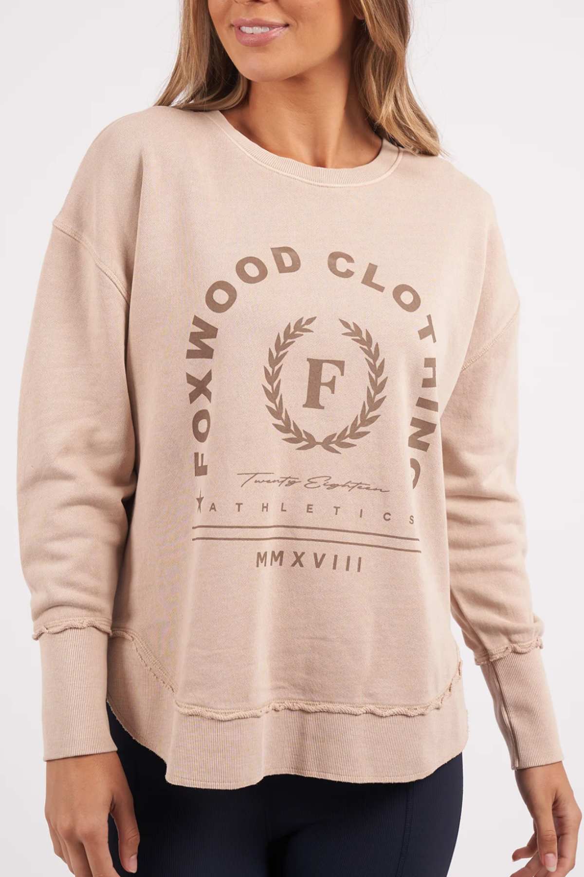 A person stands facing slightly to the side, wearing the beige Foxwood Medalion Crew sweatshirt. The front of this 100% cotton fabric garment is emblazoned with "FOXWOOD CLOTHING" and the Roman numerals "MMXXVIII," as well as a laurel wreath and an "F" emblem in the center. It also features distinctive raw edge detailing.