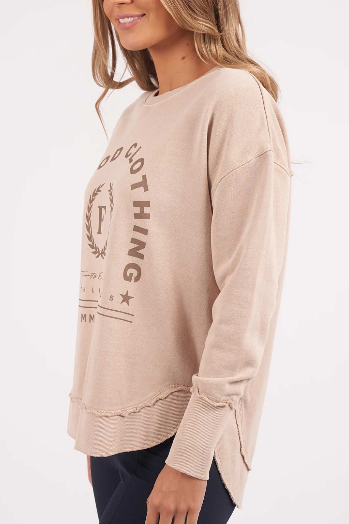 A person is wearing the Foxwood Medalion Crew, a beige long-sleeve sweatshirt featuring a logo and text on the front. Crafted from 100% cotton fabric, it offers raw edge details and side slits at the hem for a relaxed fit. The person pairs it with black pants against a plain white background.