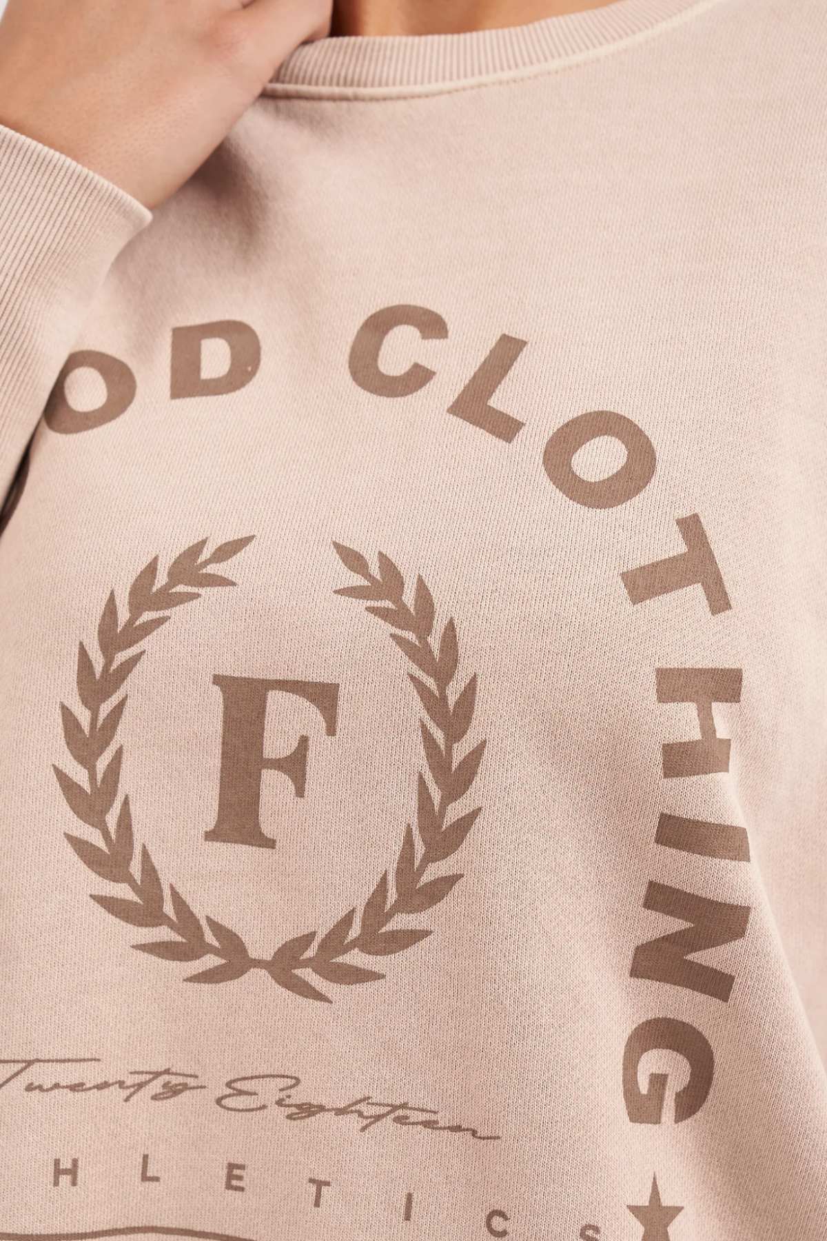 Close-up of a person wearing the Foxwood Medalion Crew, a beige sweatshirt made from 100% cotton. It features the text "Food Clothing" in a circular design and the letter "F" inside a laurel wreath. Below the laurel wreath, there are additional texts including "Twnty Eight Athletics." The sweatshirt also has raw edge detail for added style.