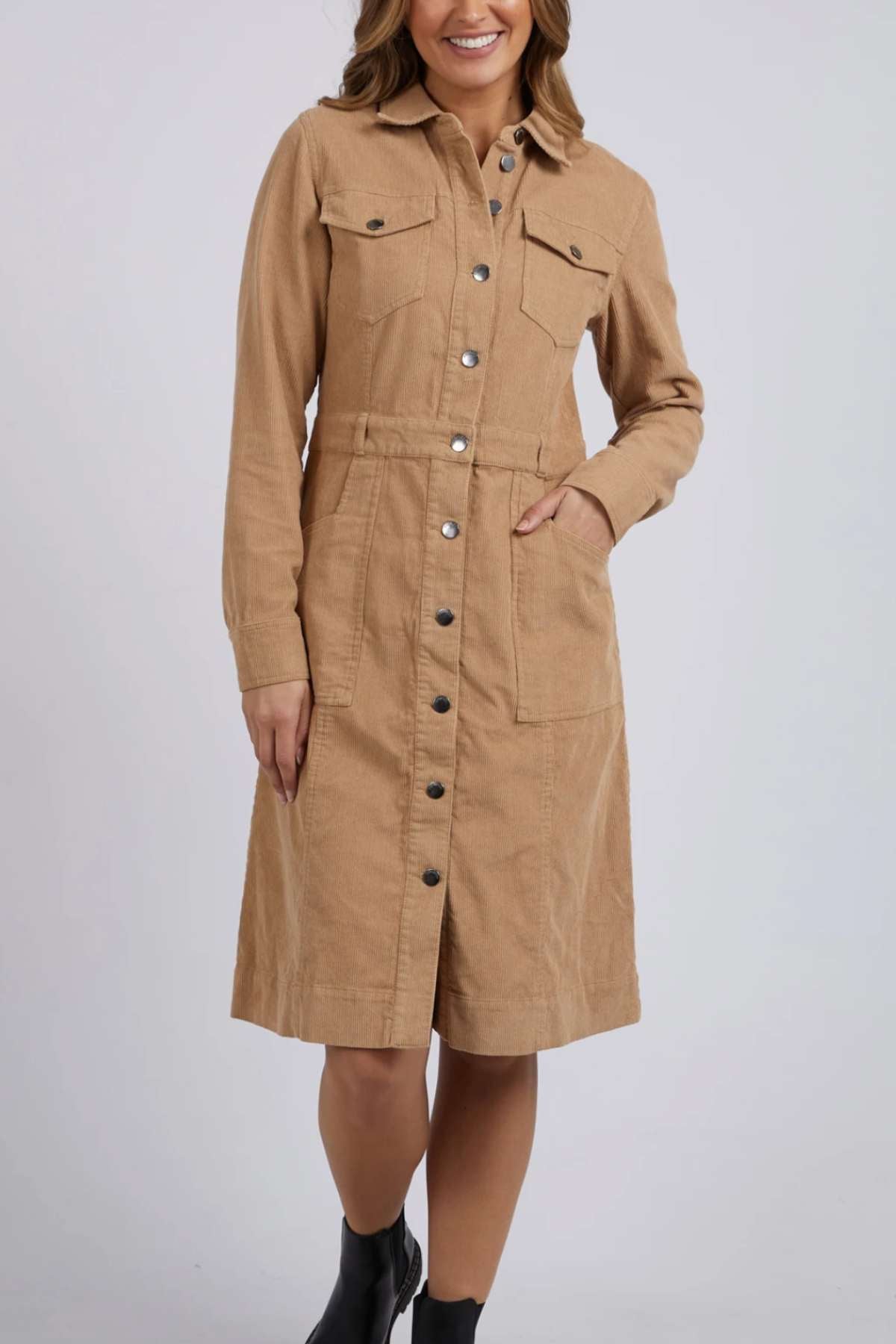 A woman wearing the Foxwood Montana Cord Dress—a knee-length, 100% cotton cord dress with long sleeves and chest pockets—paired with black ankle boots, stands against a plain light background.