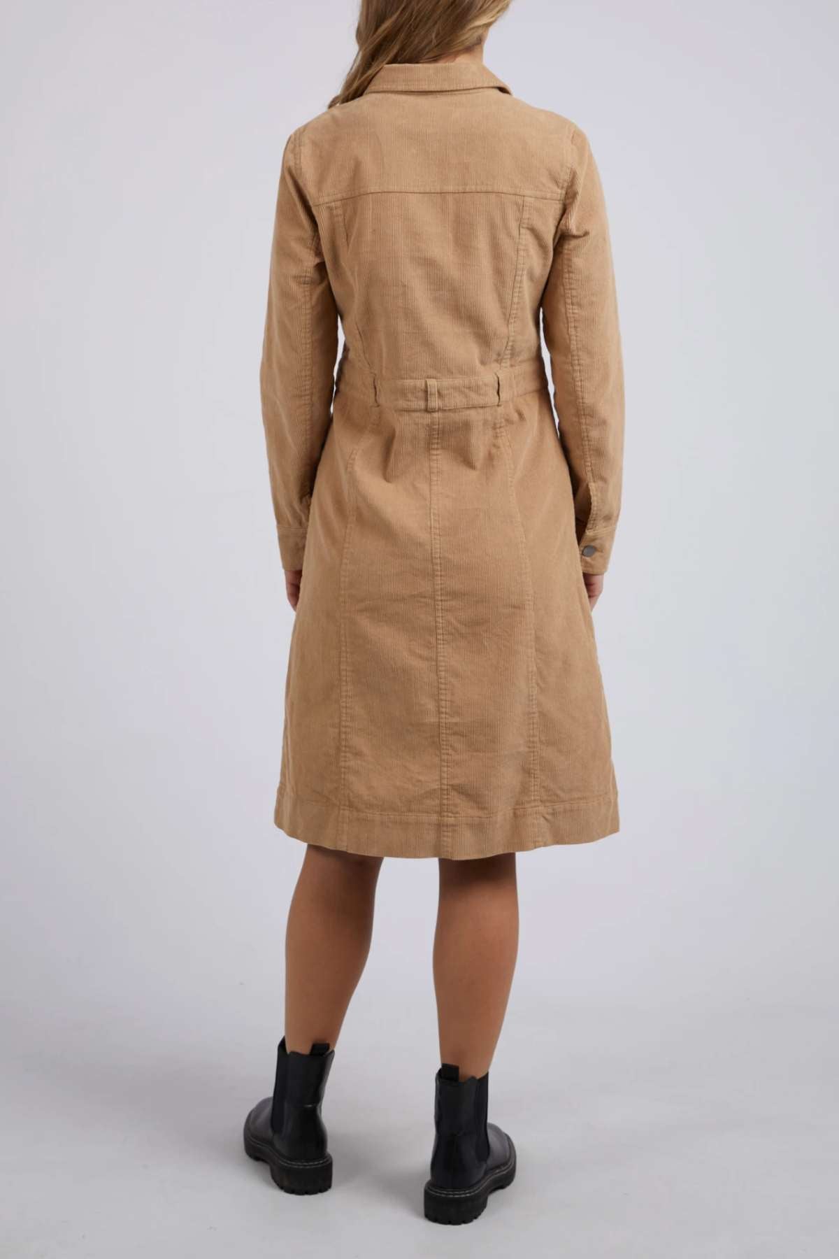 A woman faces away, wearing the Foxwood Montana Cord Dress—knee-length, 100% cotton with a belted waist—paired with black ankle boots against a plain light background.