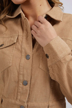 A person wearing the tan Foxwood Montana Cord Dress made of 100% cotton with metallic buttons holds the collar, with the image focusing on the upper torso to showcase the garment’s rich corduroy texture and detailed fabric.