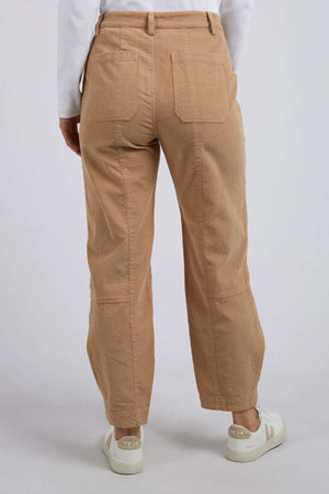 A person in relaxed-fit, tan Foxwood Montana Cord Pant made of 100% cotton is viewed from the back. They wear a white long-sleeve shirt and white Veja sneakers, standing against a gray background. The pants feature large back pockets.