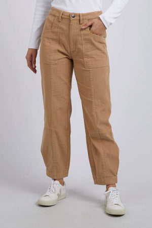Wearing high-waisted, tan Foxwood Montana Cord Pants crafted from 100% cotton and white sneakers, the person pairs them with a white long-sleeve top. One hand rests by their side against a plain gray background.