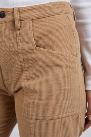 A close-up shows a person wearing Foxwood Montana Cord Pant in tan, made from 100% cotton. The relaxed fit emphasizes the waist and pockets, displaying the ribbed fabric texture alongside a crisp white long-sleeve top.