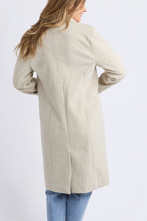 Woman with light brown hair stands with her back to the camera, wearing blue jeans and the mid-length Foxwood Nelson Coat in light beige against a plain light background.