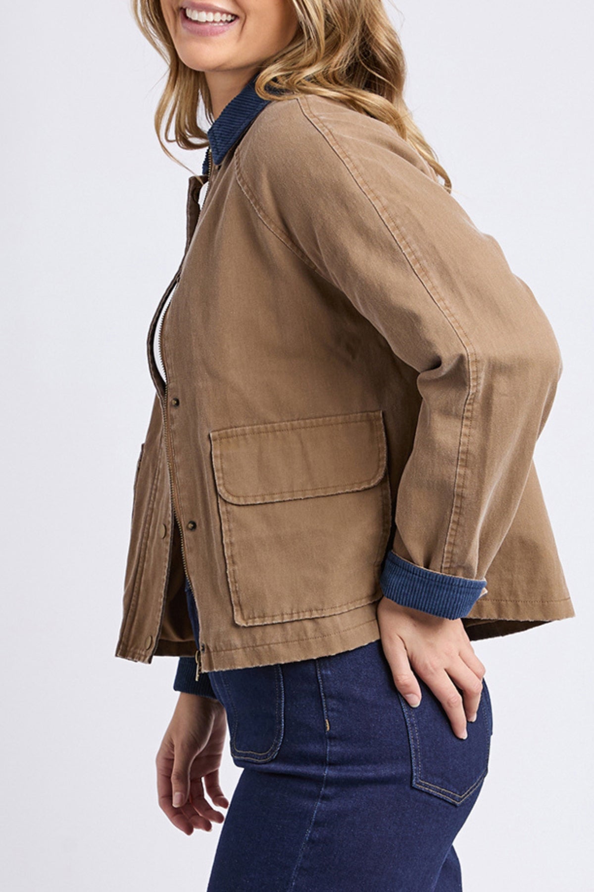 A woman smiles and poses in the Foxwood Colwood Jacket, featuring large front pockets and a contrast collar, layered over a blue collared shirt and dark blue jeans against a plain light background.