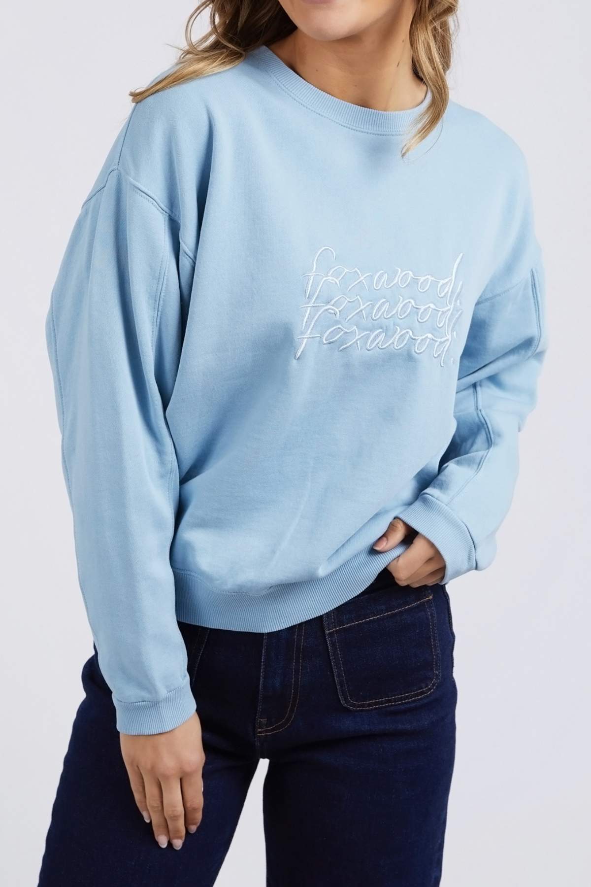 A person wearing a Foxwood Nimble Crew sweatshirt in light blue, featuring white stylized cursive writing repeated vertically three times, and dark blue jeans stands against a plain background. The relaxed fit top is made from 100% cotton.