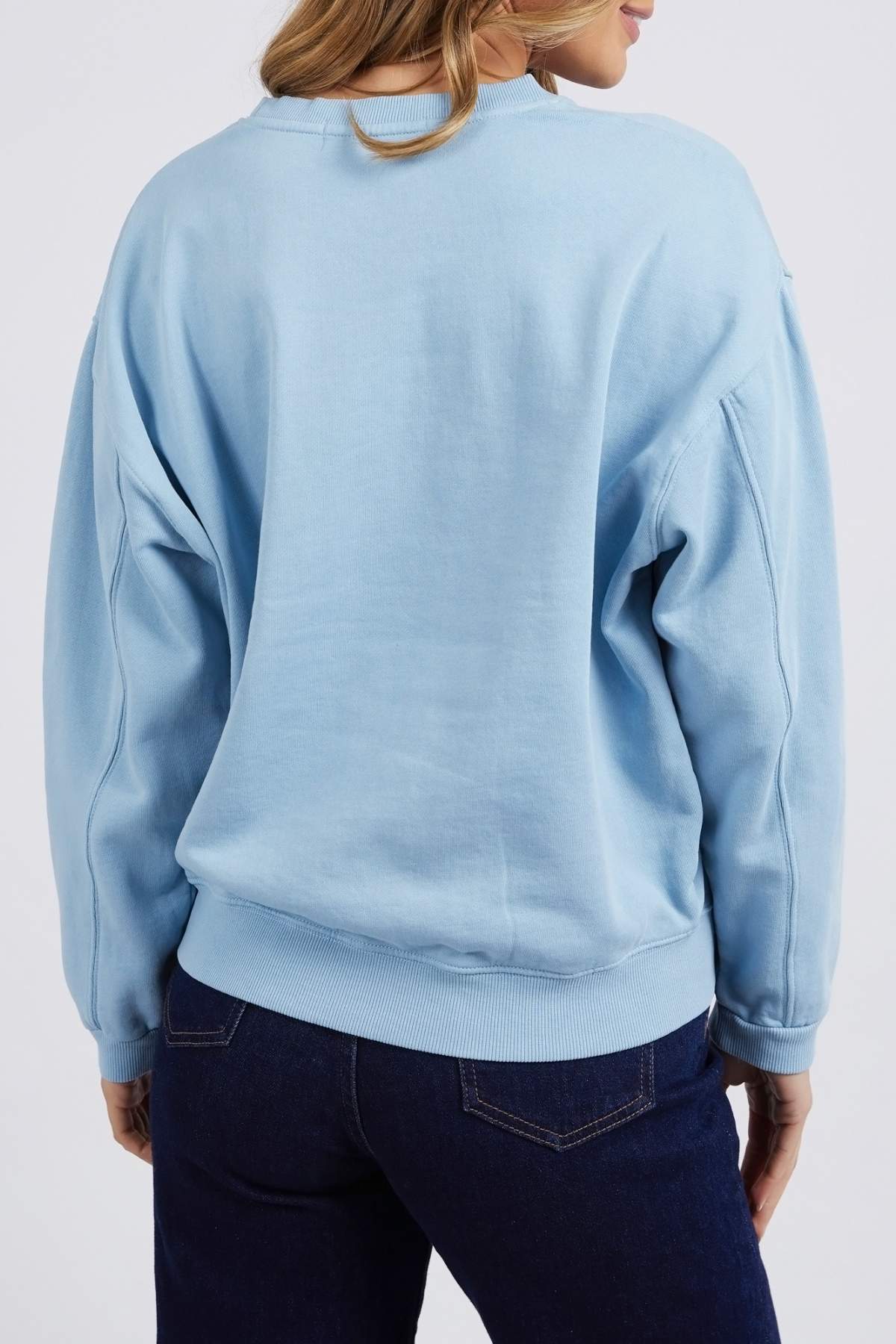A person with long hair, facing away from the camera, is dressed in a light blue Foxwood Nimble Crew sweatshirt paired with dark blue jeans. The relaxed fit of the top gives it a loose and comfortable appearance, while the jeans feature visible pockets on the back. They are set against a plain white backdrop.
