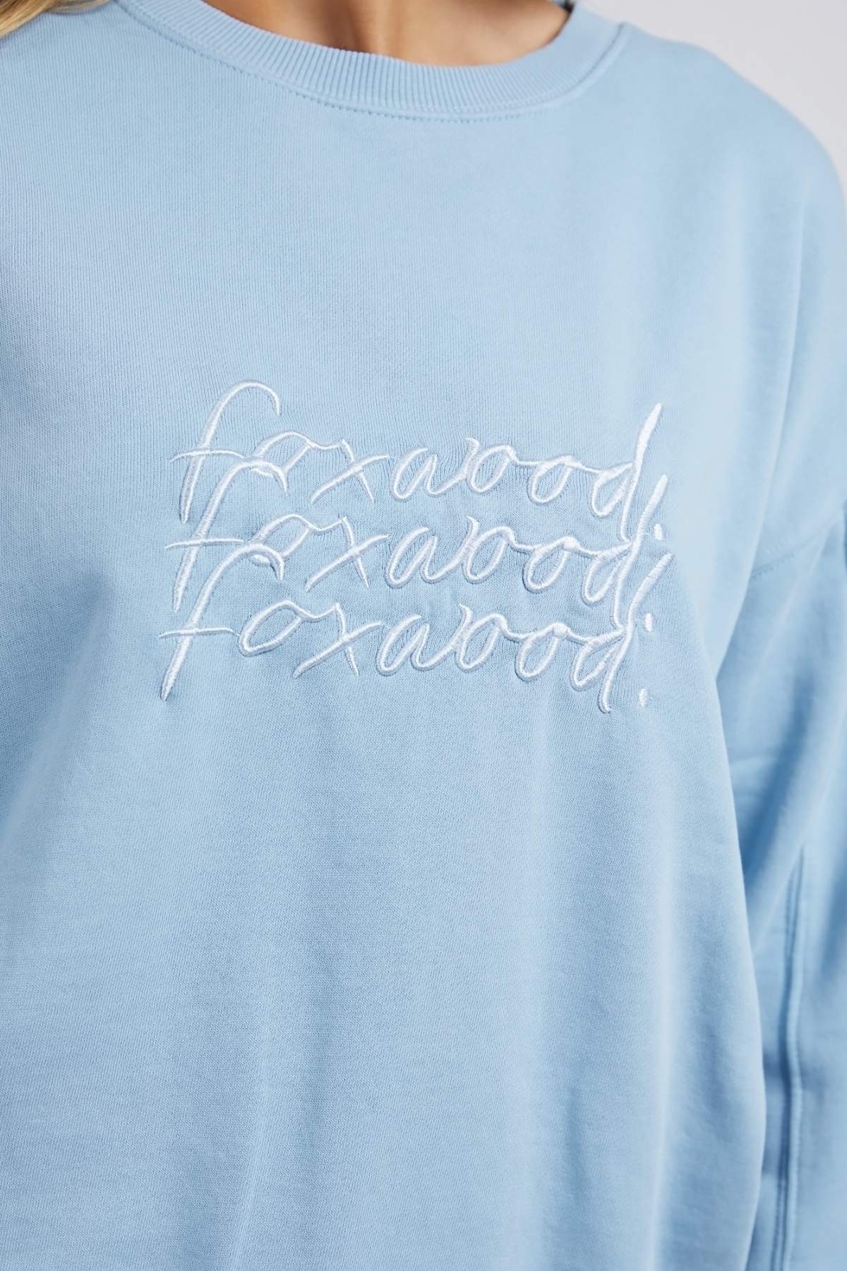 Close-up of a person wearing a light blue Foxwood Nimble Crew sweatshirt with white embroidered text. Made from 100% cotton for ultimate comfort, this relaxed-fit top ensures ease of movement. The text seems to repeat and overlap, making it difficult to discern the word.