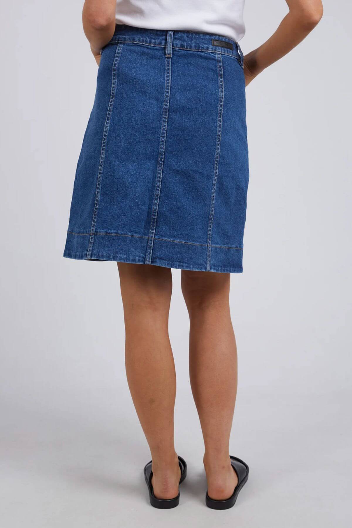 Someone stands facing away from the camera against a plain light background, wearing a blue Foxwood Nolan Skirt with contrast stitching, paired with a white top and black sandals.