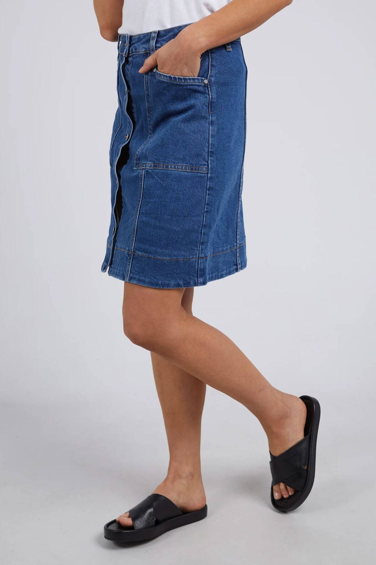 A person wears the Foxwood Nolan Skirt—a knee-length blue skirt featuring front buttons, large pockets, and contrast stitching—paired with a white top and black slide sandals, standing before a plain light background.
