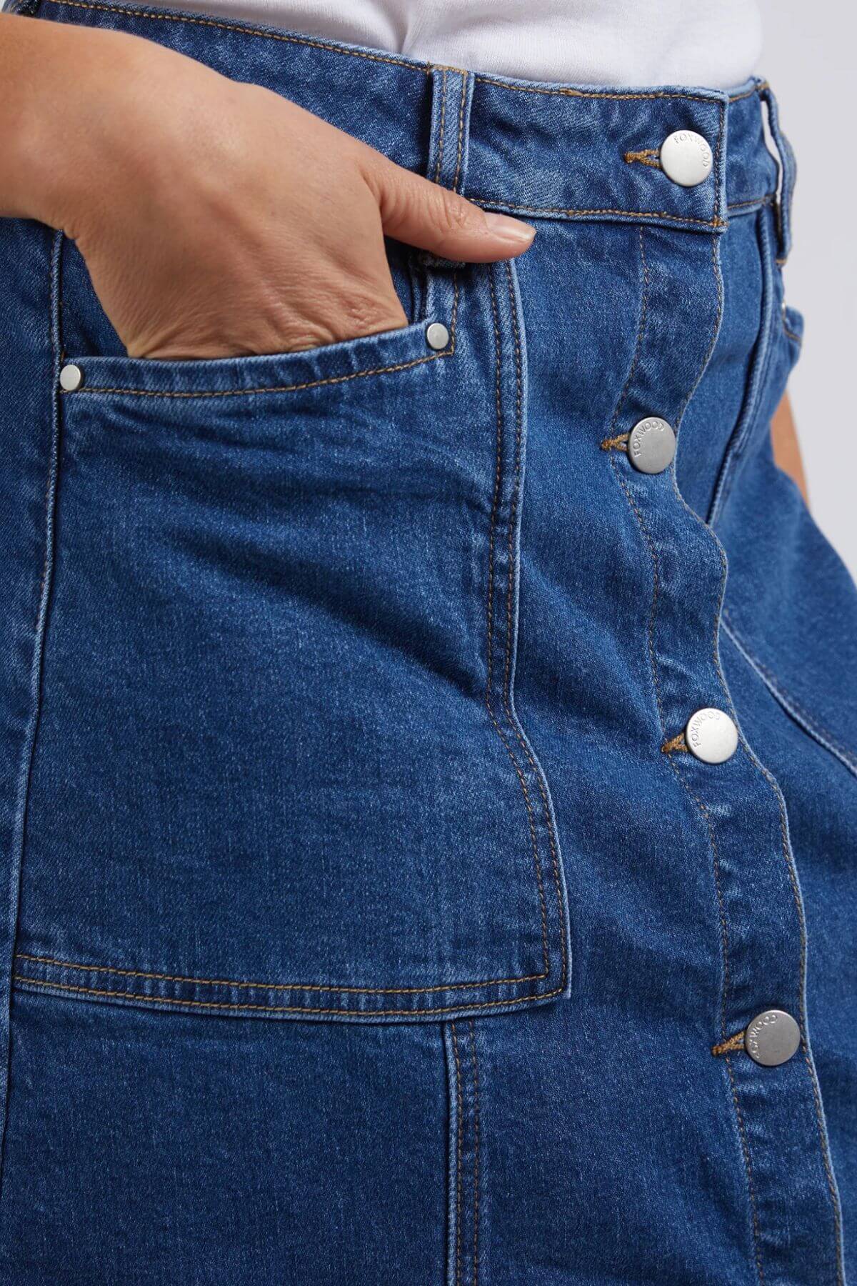 Close-up of a person wearing the Foxwood Nolan Skirt featuring metallic buttons, contrast stitching, and a large front pocket, styled casually with a white top and their hand resting inside the pocket.