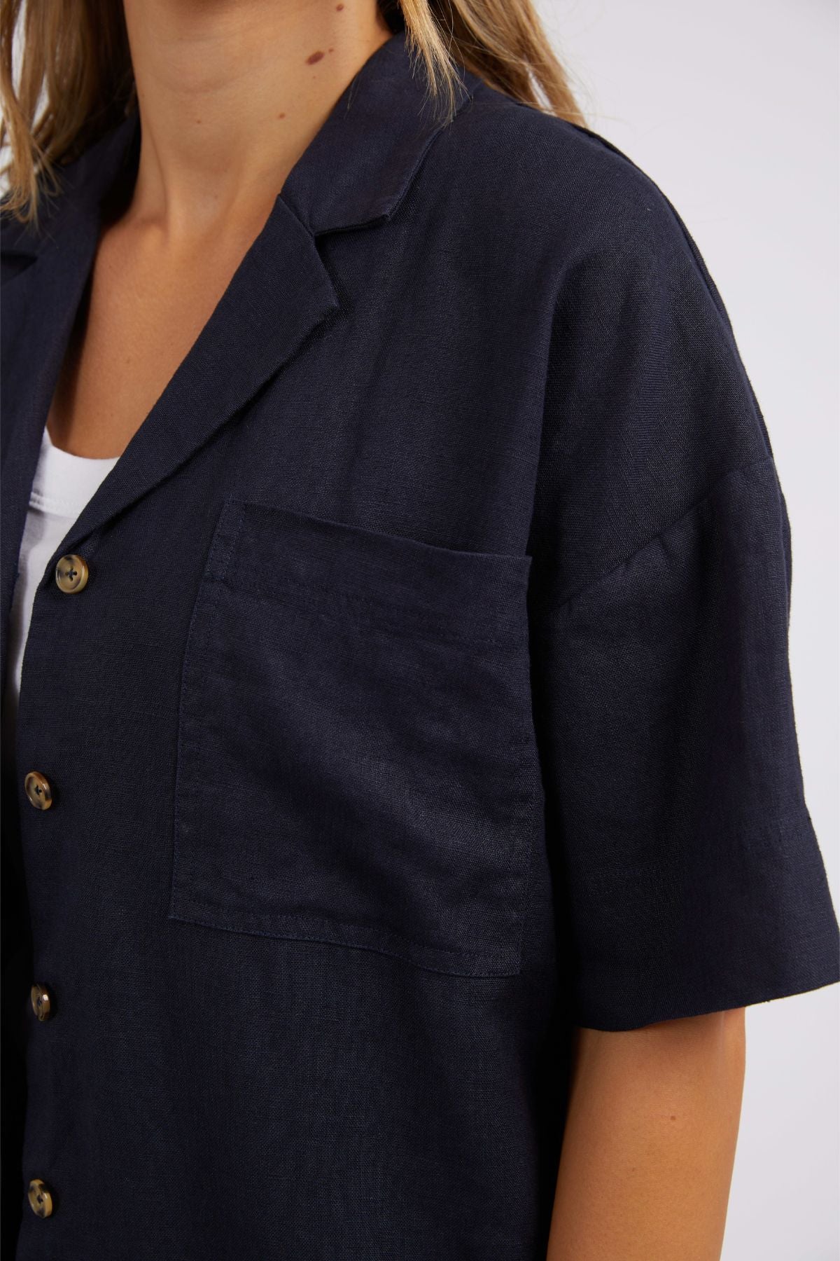 A person wears the Foxwood Oakleigh Shirt in dark navy linen, featuring a relaxed fit, large chest pocket, and notched collar layered over a white top. This short-sleeve button-up is made for comfort. Only the upper body is shown.