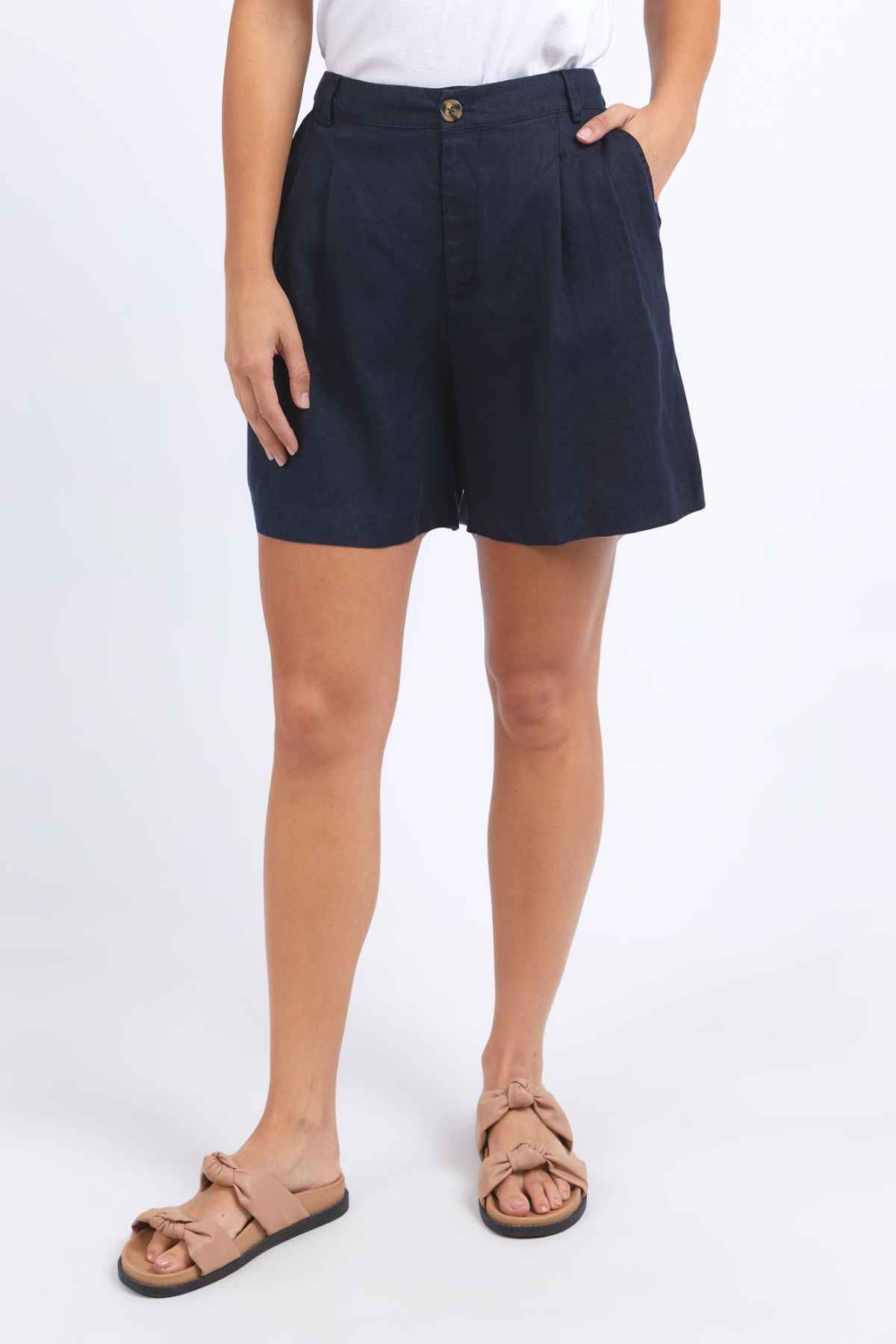 A person wearing Foxwood Oakleigh Shorts, paired with a white shirt, stands against a plain background. They complete the look with beige sandals featuring bows.