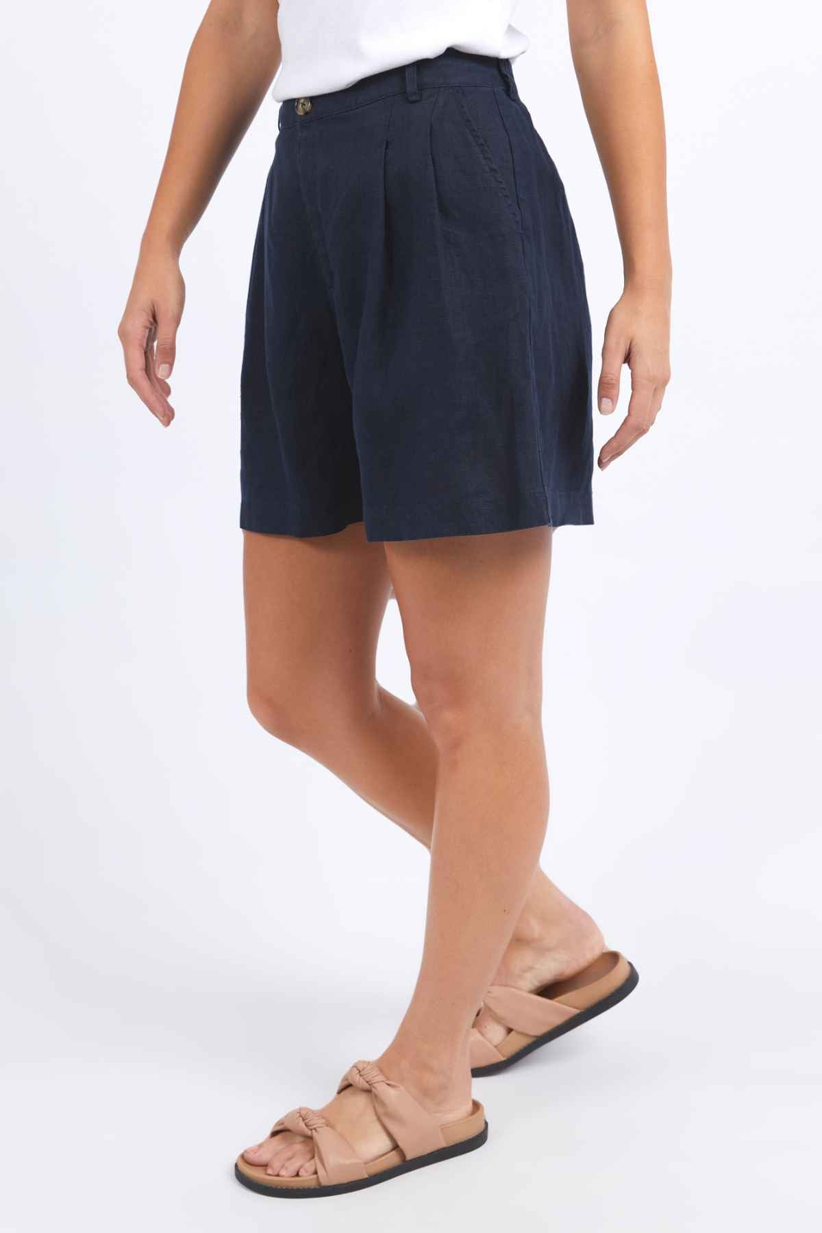 A person is wearing a white top, navy Foxwood Oakleigh Shorts with a button detail, and nude slip-on sandals featuring a bow. They are standing against a plain white background.