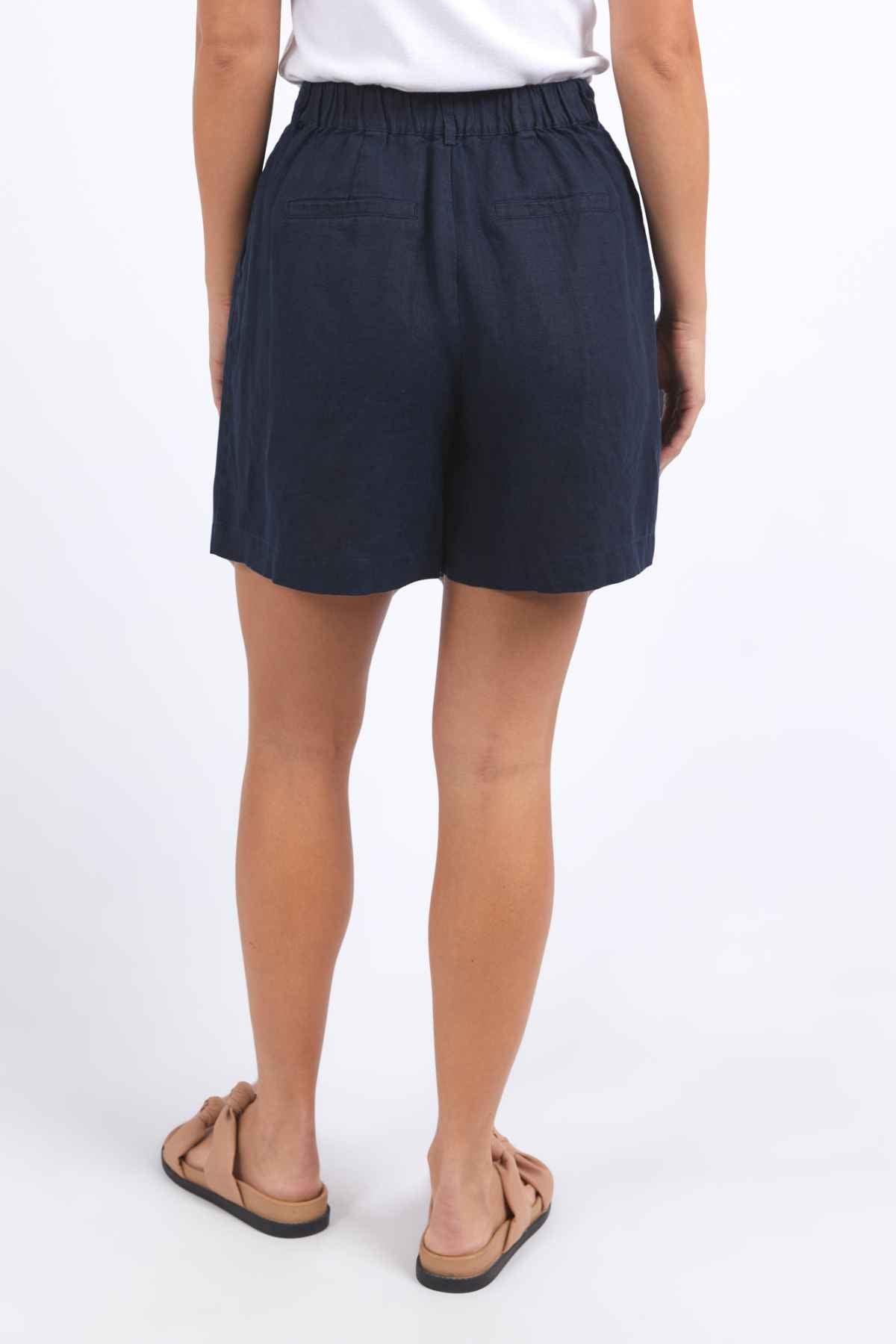 A person wearing a white top and navy blue Foxwood Oakleigh Short, featuring an elastic waistband, stands facing away from the camera. They are wearing light brown sandals, all against a plain white backdrop.