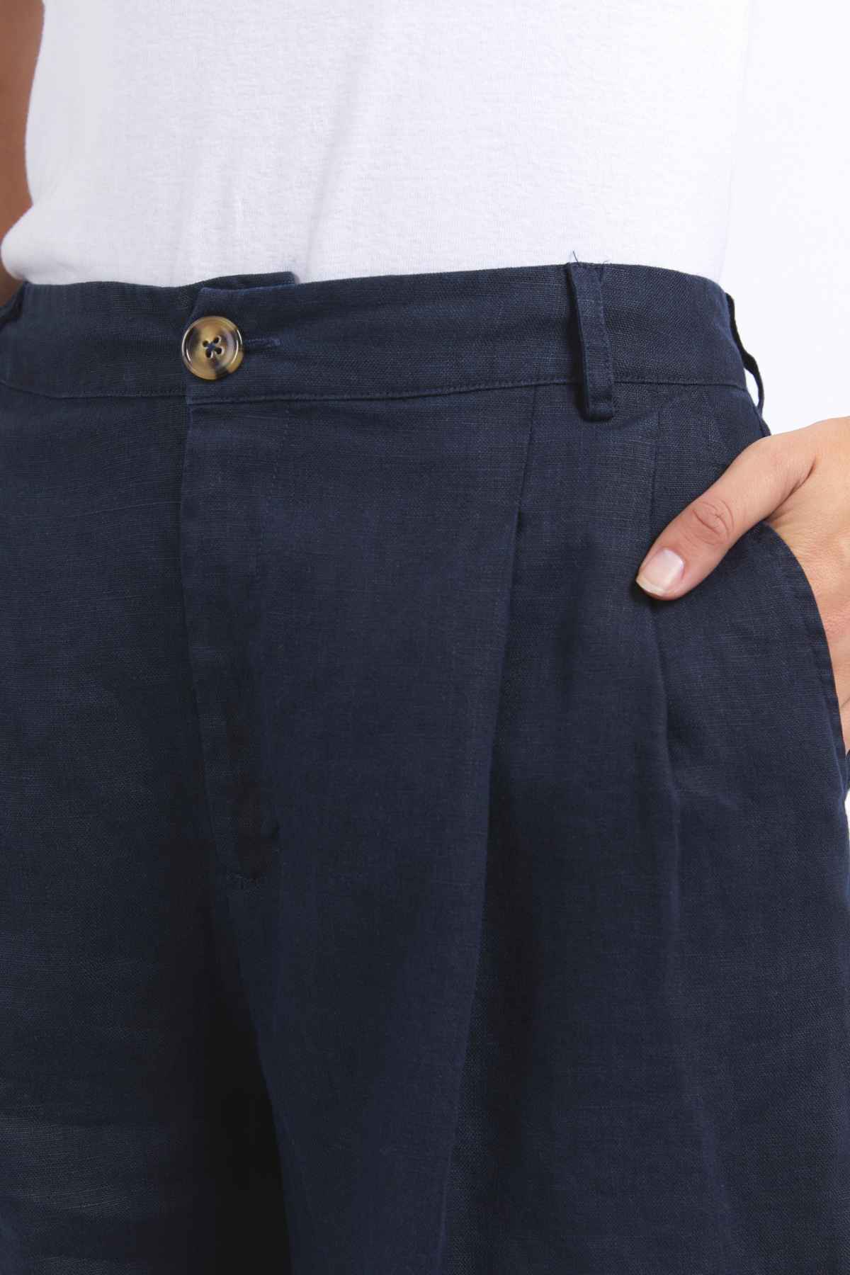 A person is wearing dark blue pants featuring a button and zipper closure. Their hand is tucked into the right pocket, and the waistband has belt loops. The pants, reminiscent of the casual comfort found in Foxwood Oakleigh Shorts, are paired with a white top.